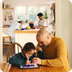 A father sits with his daughter at the kitchen table and helps her use a tablet.